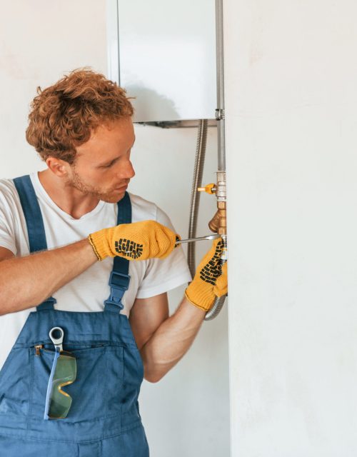 Repairing water heater. Young man working in uniform at construction at daytime.